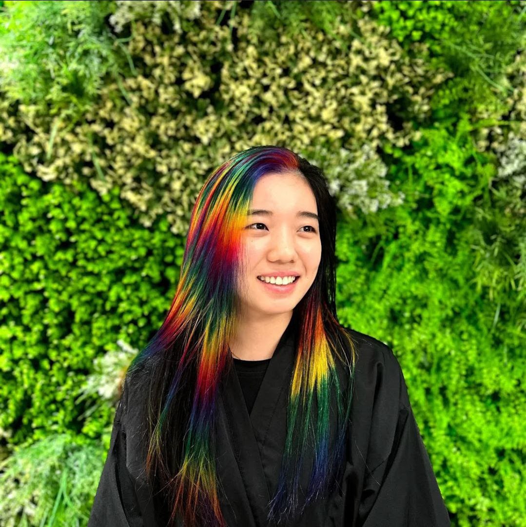 A smiling woman showcases vibrant rainbow-colored hair against a lush green wall.