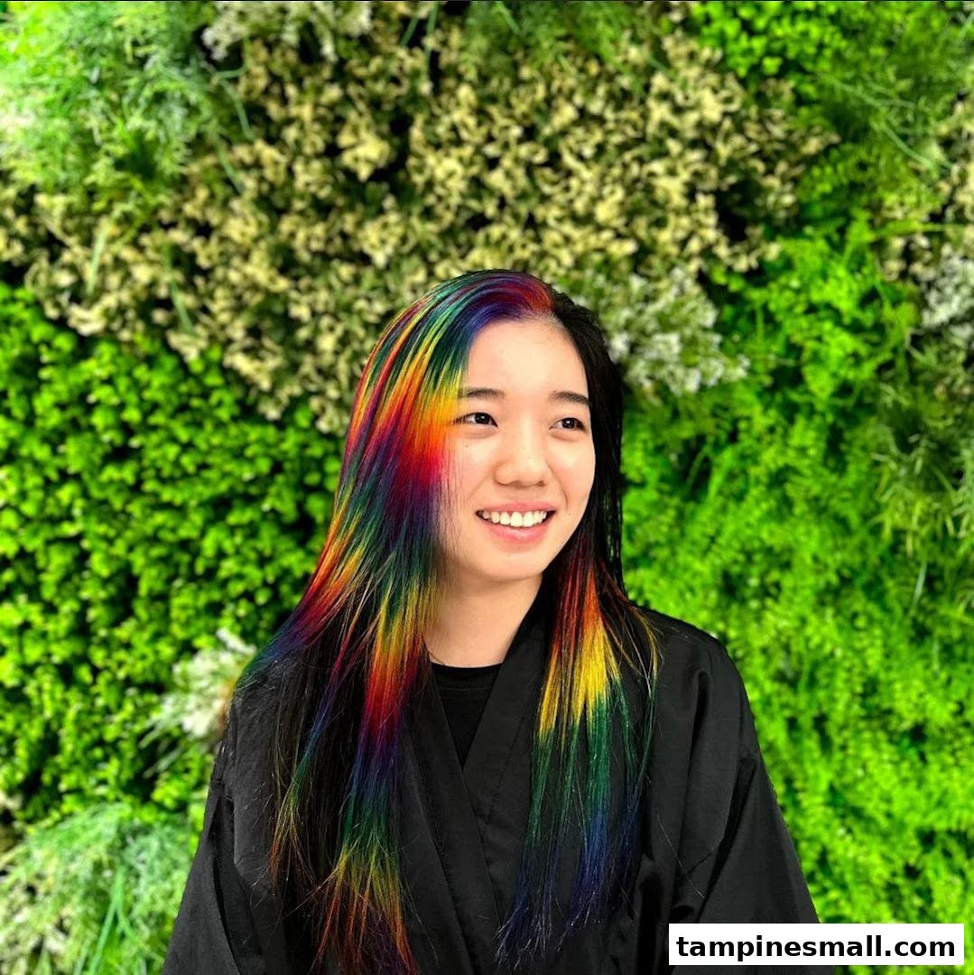 A smiling woman showcases vibrant rainbow-colored hair against a lush green wall.