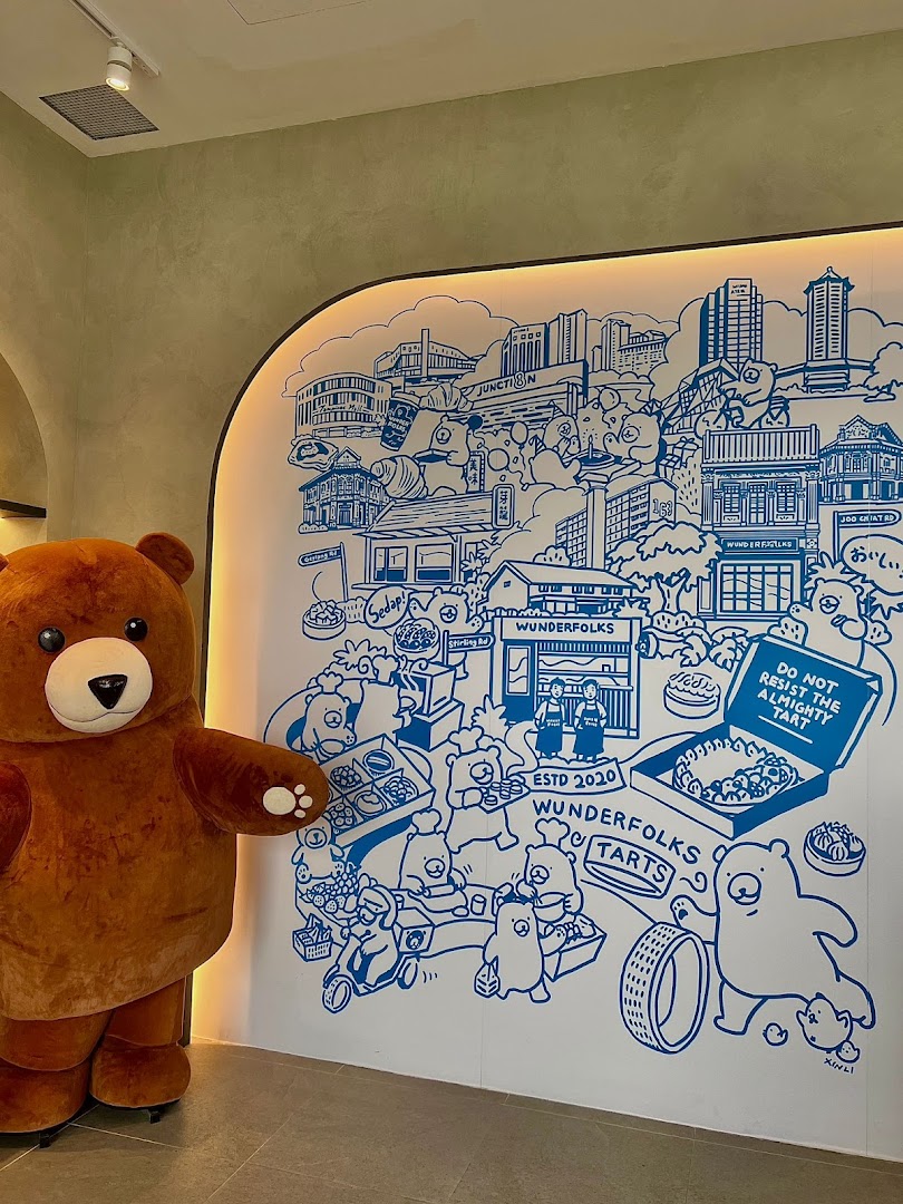 A large bear mascot stands beside a whimsical blue and white mural depicting cityscapes and bear characters.