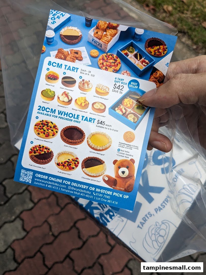 A hand holds a flyer showcasing various tarts and pastries from Wunderfolks, including pricing and ordering information.