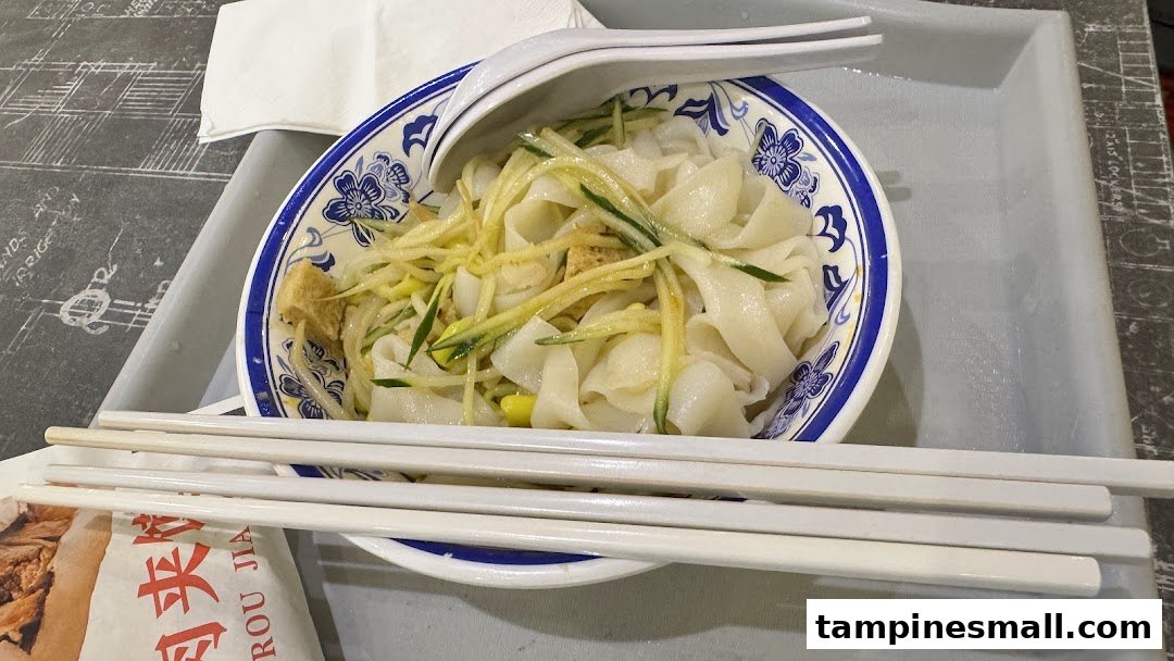 A delicious bowl of wide noodles with cucumber and tofu, served with chopsticks and spoon.