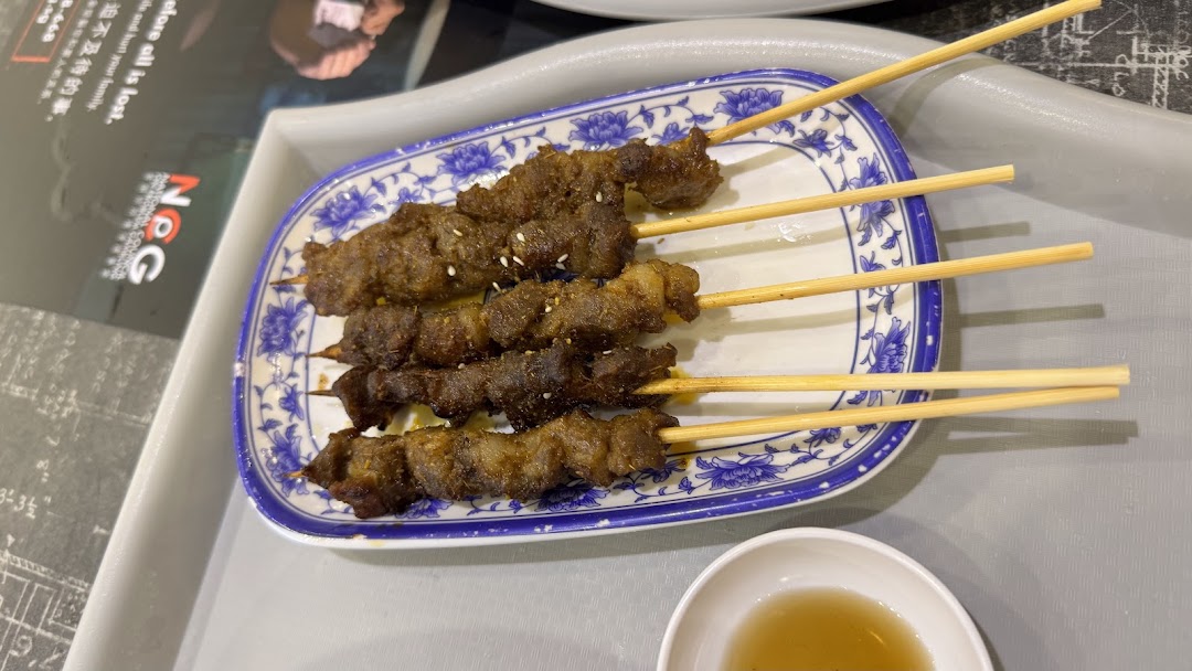 A plate of five grilled meat skewers with sesame seeds, served with a dipping sauce.