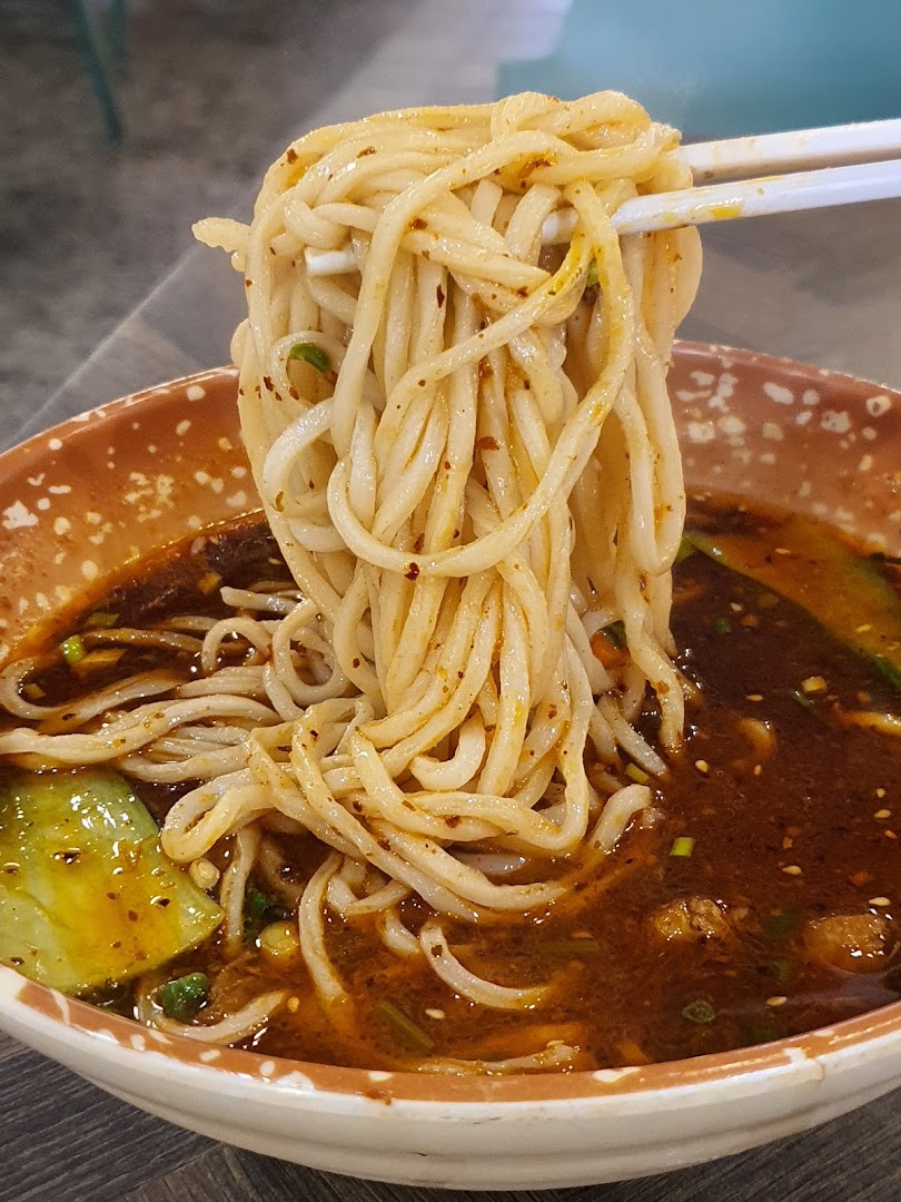Close-up of chopsticks lifting spicy noodles from a rich, dark broth with vegetables.