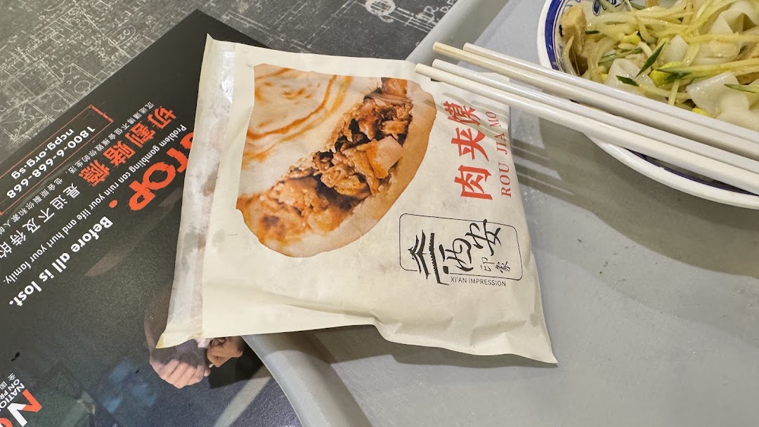 A Rou Jia Mo in a branded wrapper and a bowl of noodles on a tray, showcasing Xi'an Impression's cuisine.
