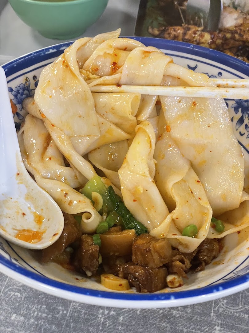 A close-up of a bowl of wide, hand-pulled noodles with braised meat and green vegetables.