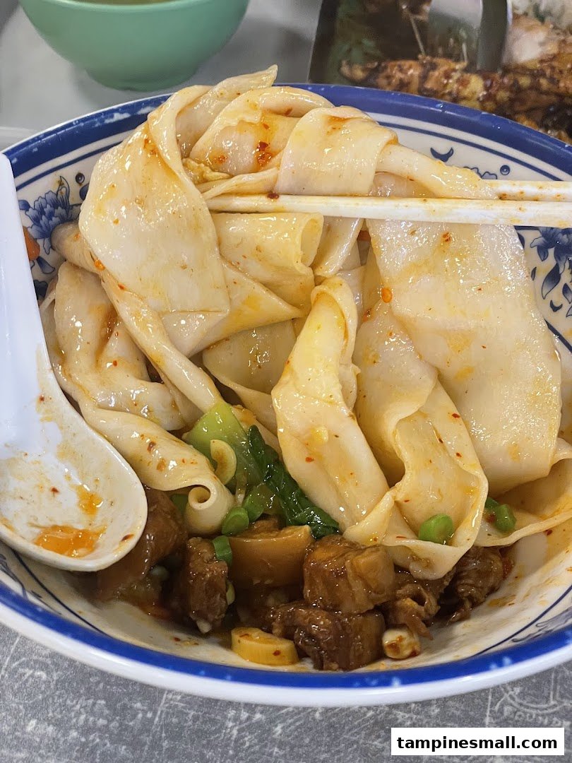 A close-up of a bowl of wide, hand-pulled noodles with braised meat and green vegetables.