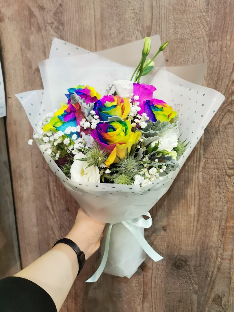 A hand holds a vibrant bouquet of rainbow roses and mixed flowers, beautifully wrapped.
