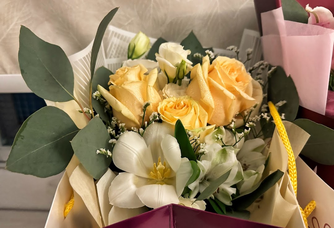 A vibrant bouquet featuring yellow roses, white tulips, and green foliage, presented in a gift bag.