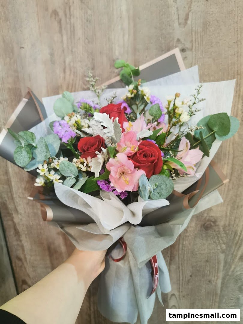 A hand holds a vibrant, beautifully wrapped floral bouquet featuring red roses and mixed blooms.