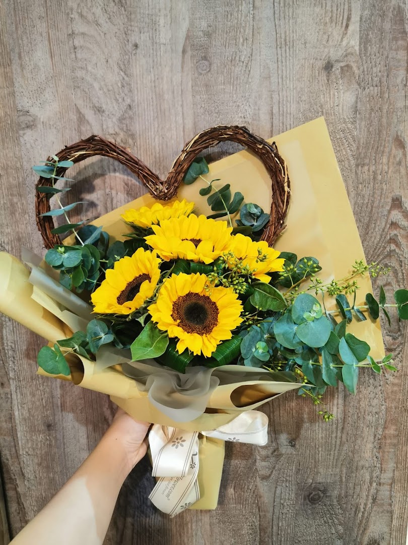 A hand holds a vibrant sunflower bouquet with eucalyptus and a heart-shaped vine accent.