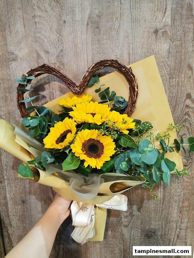 A hand holds a vibrant sunflower bouquet with eucalyptus and a heart-shaped vine accent.