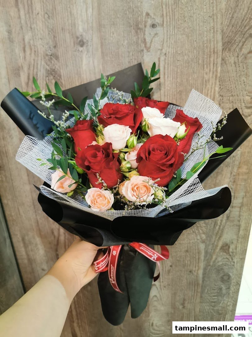 A hand holds a beautifully wrapped bouquet of red, white, and light pink roses with green foliage.