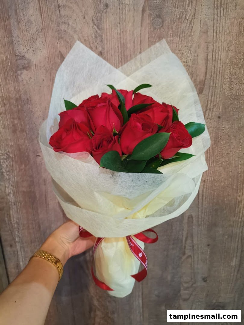 A hand holds a beautiful bouquet of vibrant red roses wrapped in delicate paper and tied with a red ribbon.