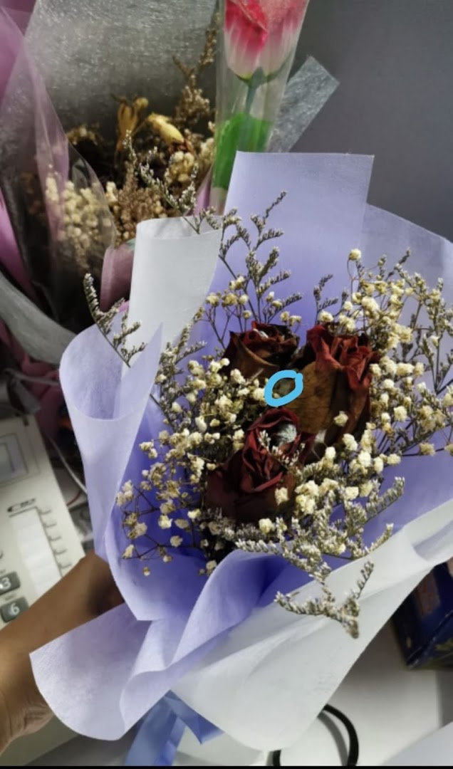 A hand holds a bouquet of dried red roses and baby's breath wrapped in purple paper.
