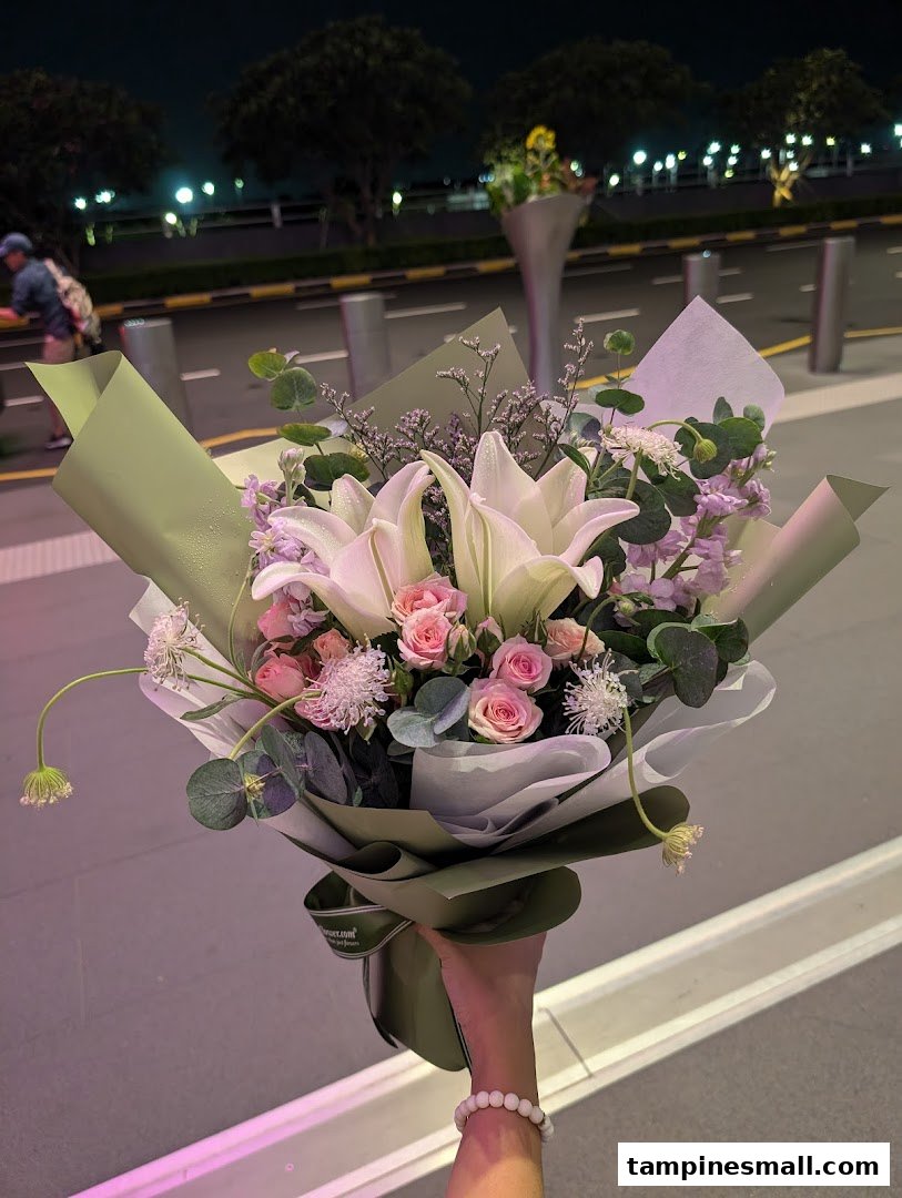 A hand holds a beautifully wrapped bouquet of fresh lilies, roses, and other flowers at night.