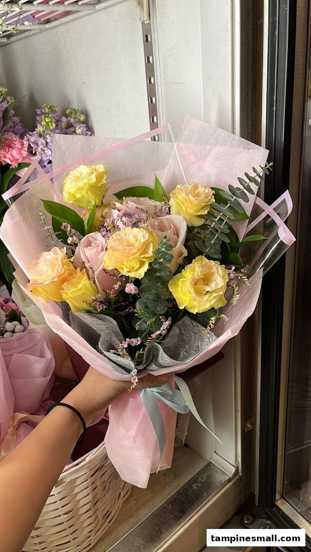 A hand holds a beautifully wrapped bouquet of yellow and pink flowers with green foliage.