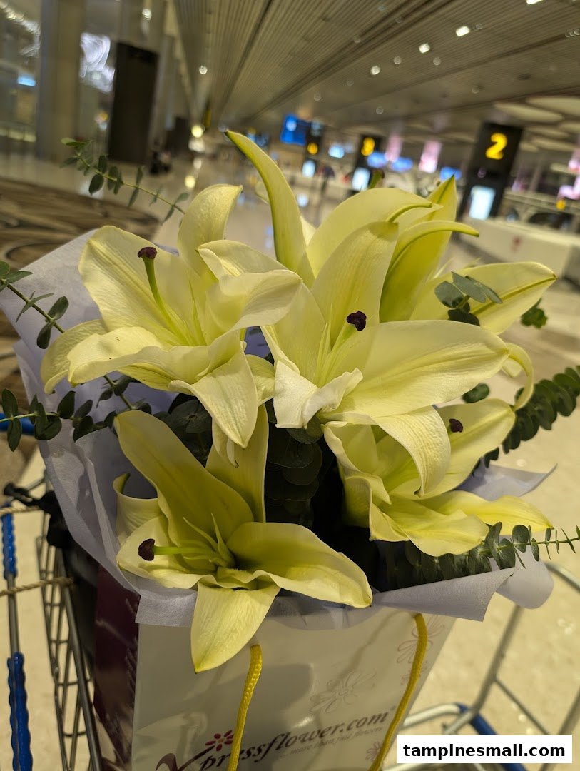 A beautiful bouquet of white lilies from Xpressflower in a shopping bag, set against a blurred airport background.
