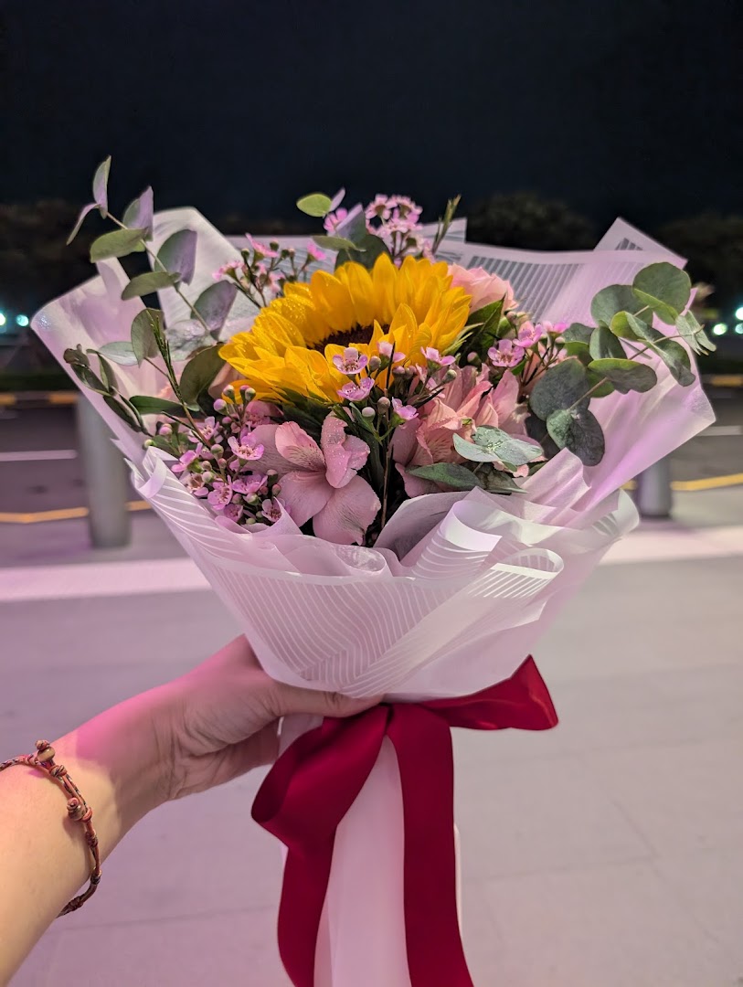 A hand holds a vibrant bouquet featuring a sunflower, pink flowers, and eucalyptus, wrapped in white paper with a red ribbon.