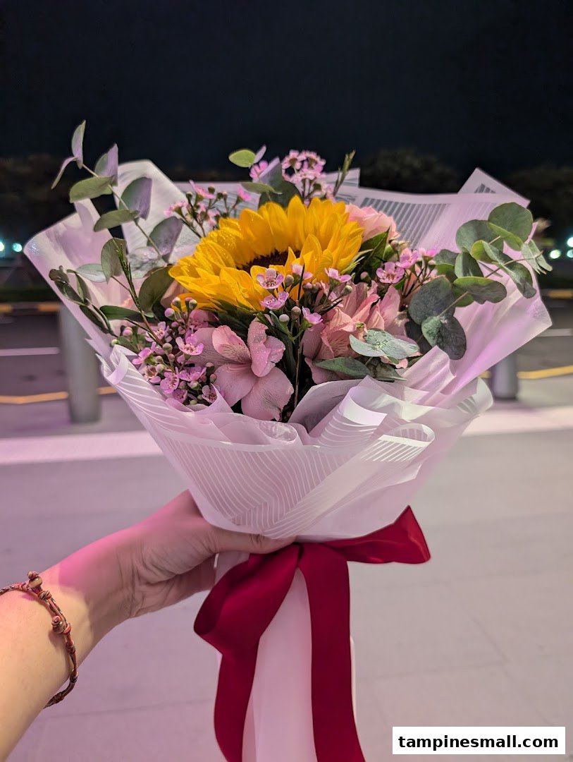 A hand holds a vibrant bouquet featuring a sunflower, pink flowers, and eucalyptus, wrapped in white paper with a red ribbon.