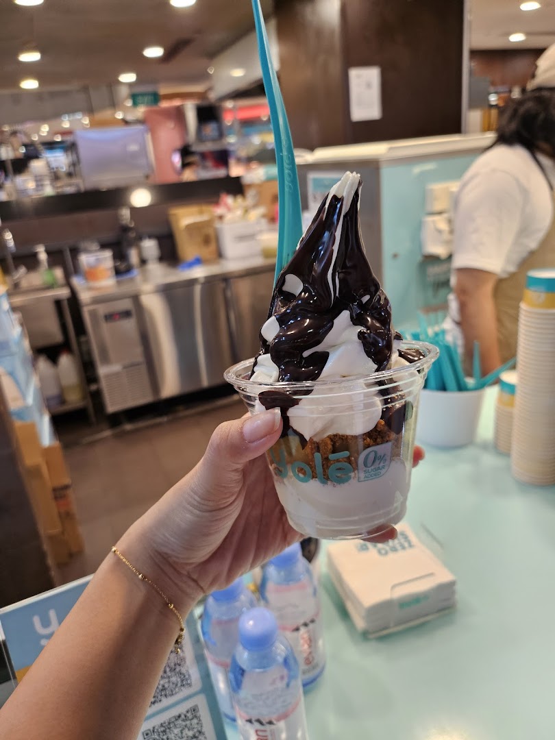 A hand holding a Yolé frozen yogurt cup with chocolate sauce and toppings inside the shop.
