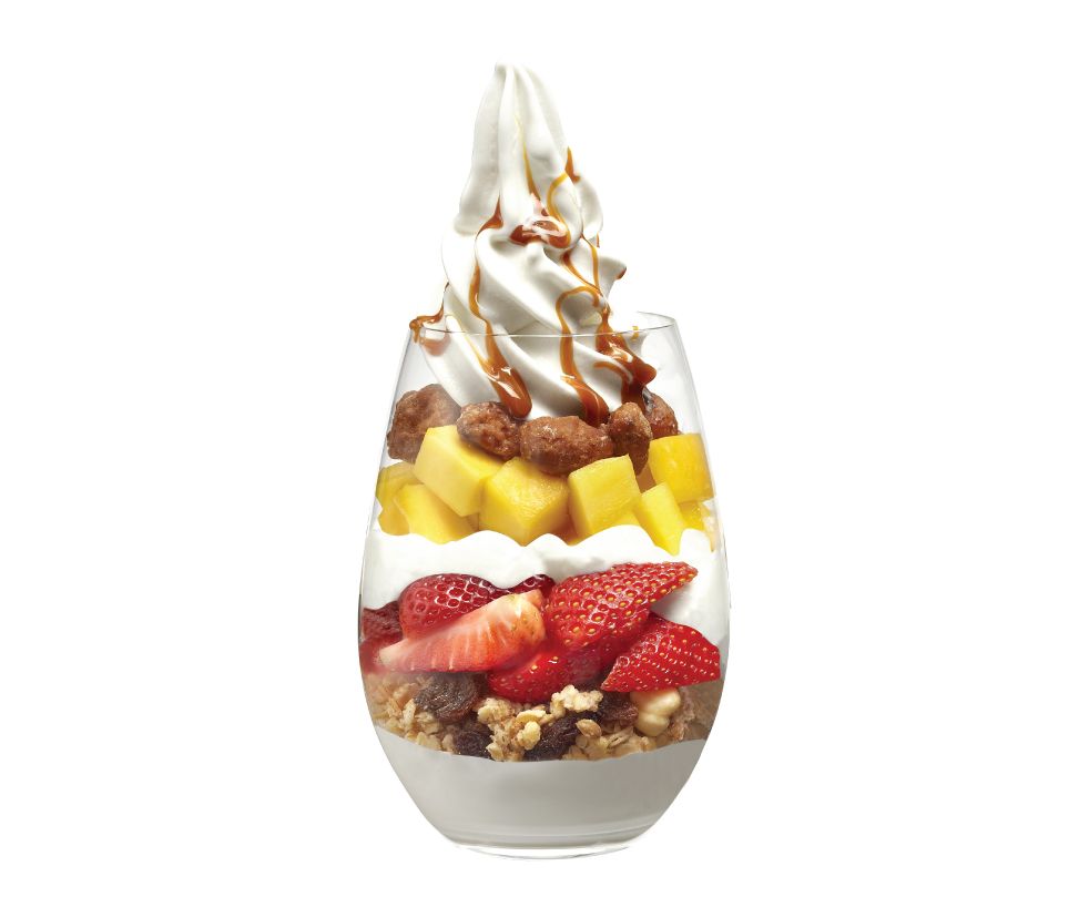 A vibrant layered dessert featuring frozen yogurt, fresh strawberries, mango, granola, and caramel drizzle.