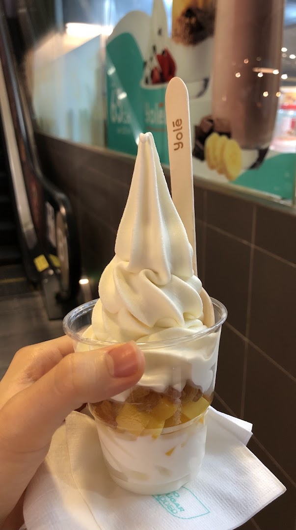 A hand holds a cup of Yolé frozen yogurt with fruit layers and a branded wooden spoon.