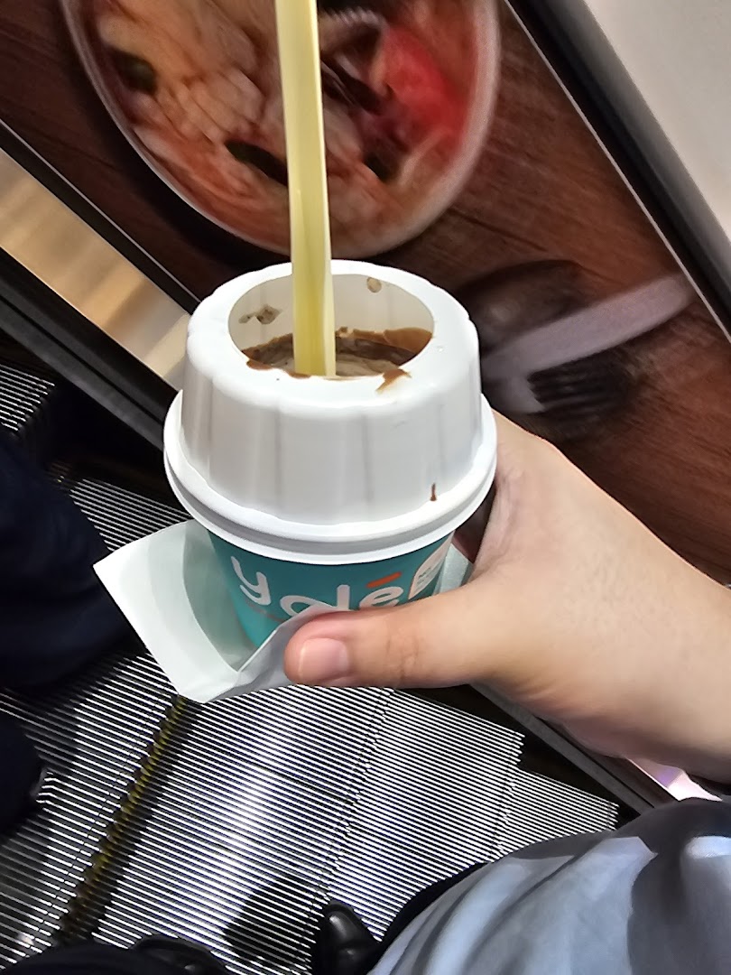 A hand holds a Yolé frozen yogurt cup with a straw while on an escalator.