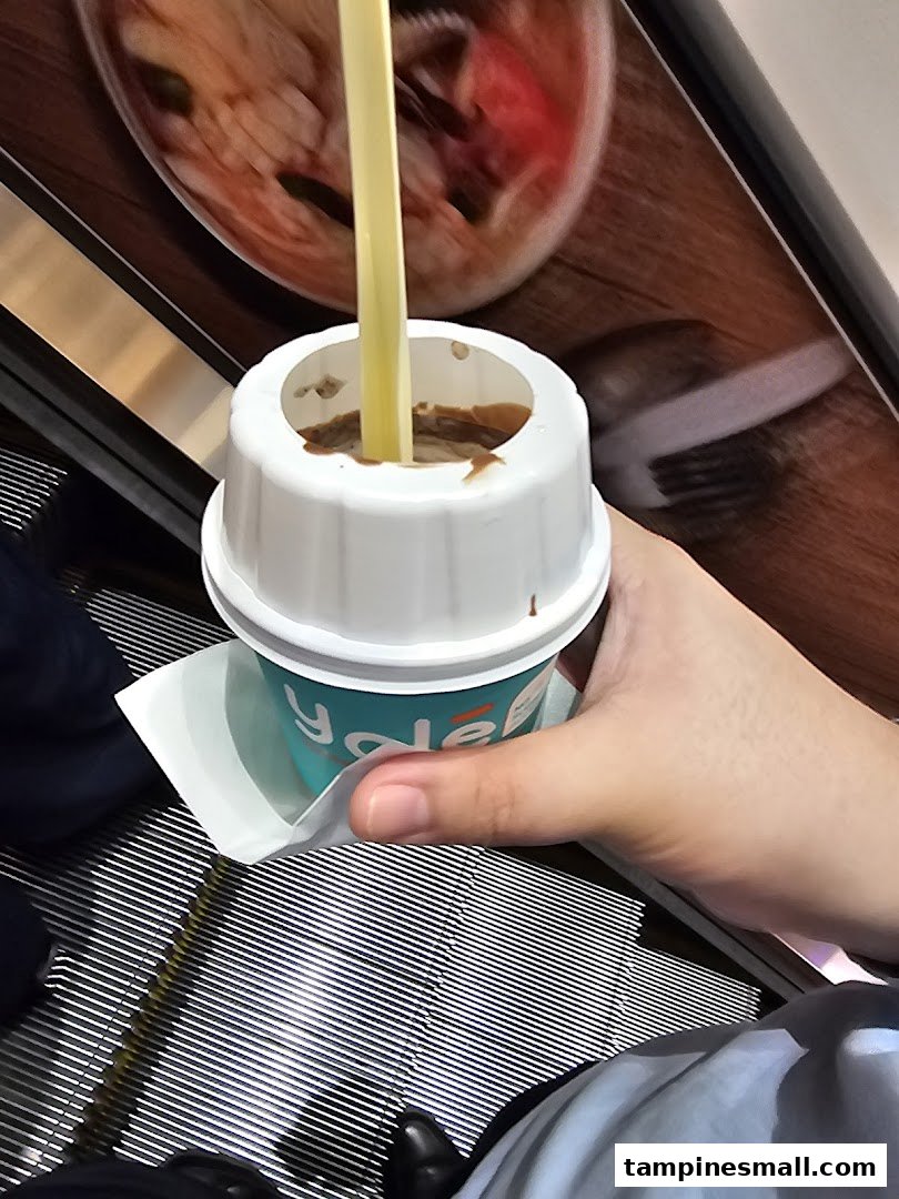 A hand holds a Yolé frozen yogurt cup with a straw while on an escalator.