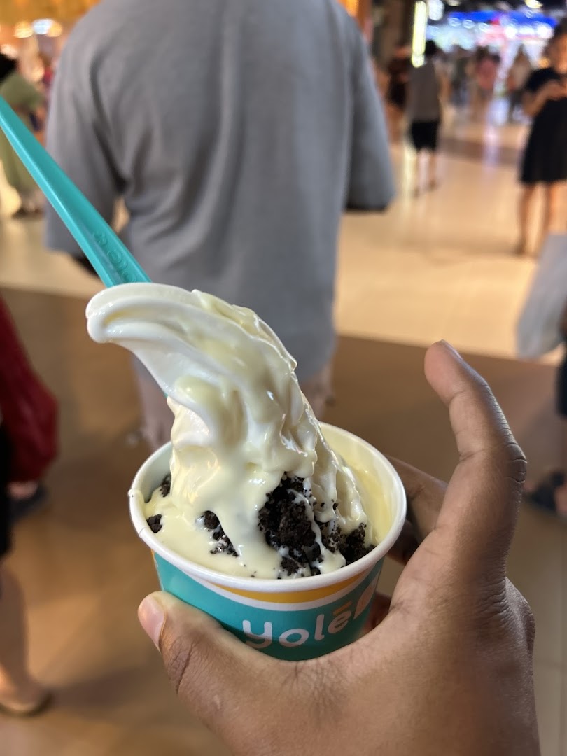 A hand holds a Yolé frozen yogurt cup with white chocolate sauce and cookie crumbs.