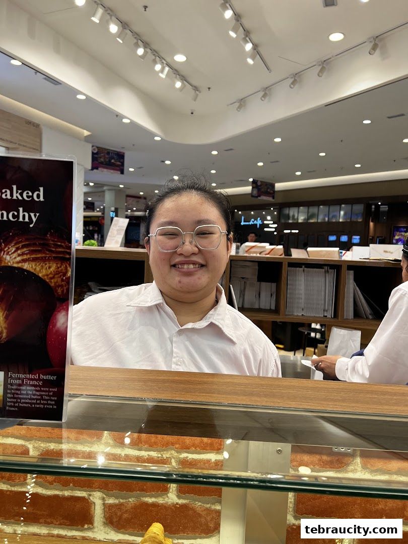 A smiling person in a white shirt stands behind a counter with baked goods and a sign.