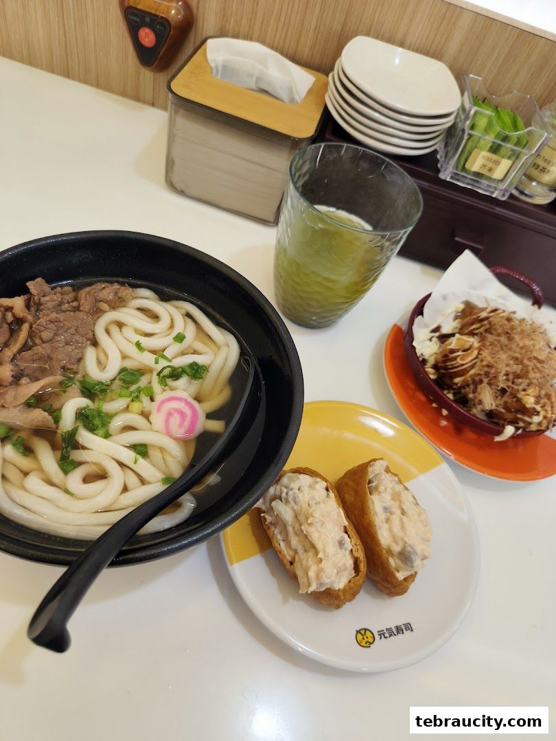 A table setting with a bowl of udon, inari sushi, takoyaki, and a glass of green tea.