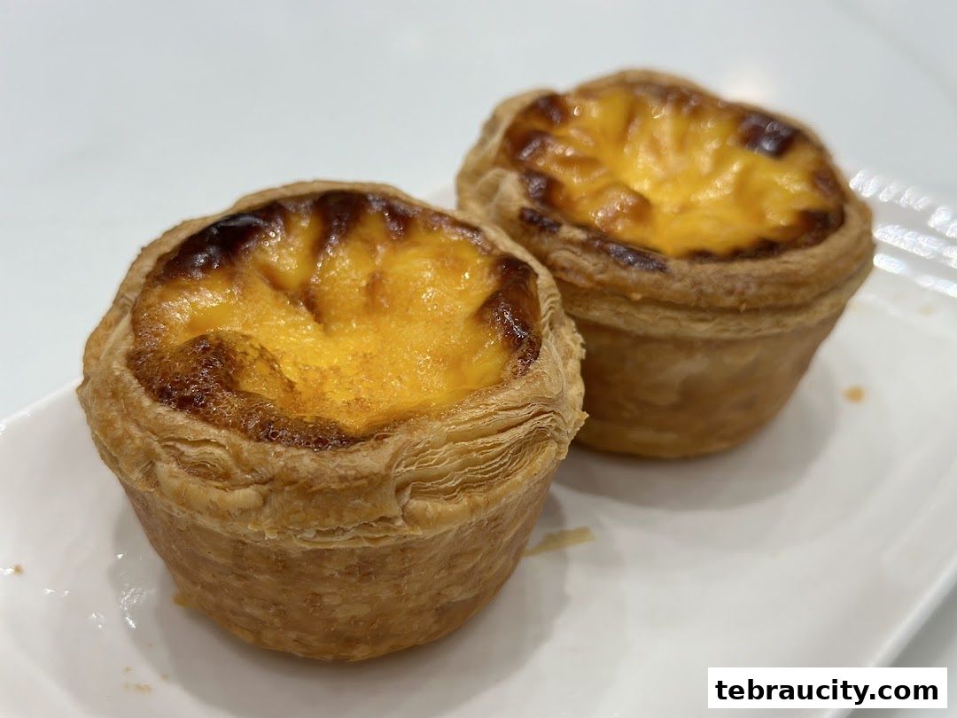 Two delicious Portuguese egg tarts with flaky pastry and caramelized tops on a white plate.