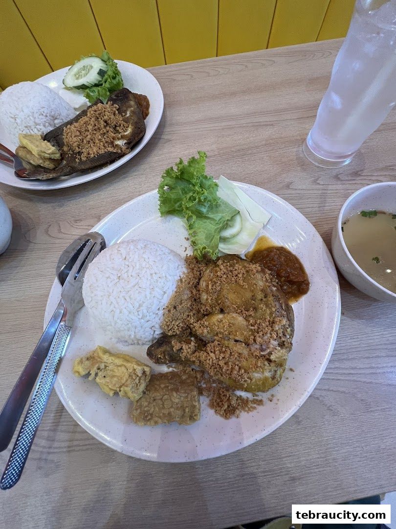 Two plates of Indonesian fried chicken with rice, tofu, and vegetables, served with soup and a drink.