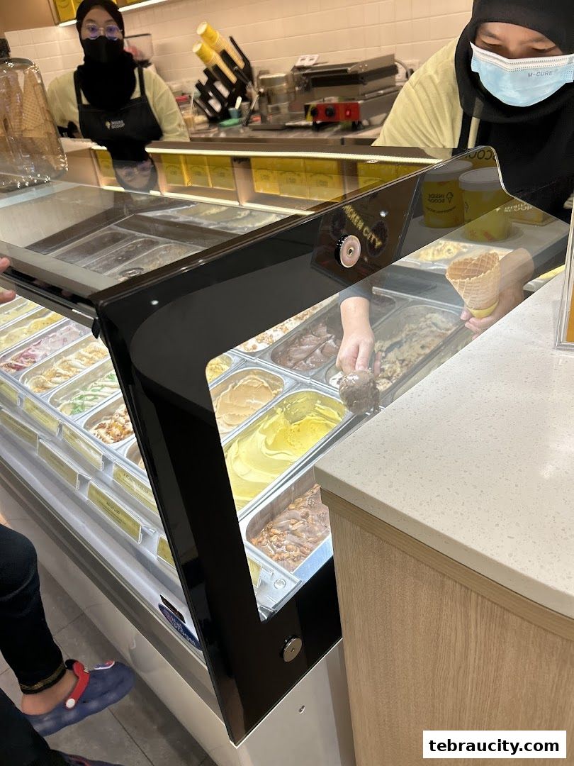 Staff serving ice cream from a display freezer to a customer holding a cone.