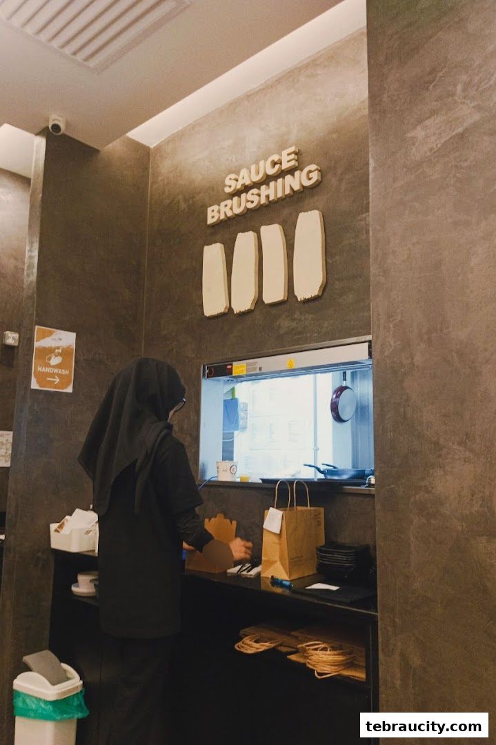A person in a hijab stands at a counter in a restaurant with a sign that says 'SAUCE BRUSHING'.