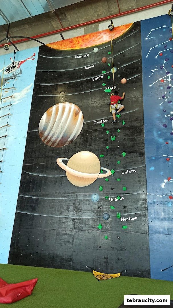 A child climbs a colorful indoor rock wall themed with planets and constellations.