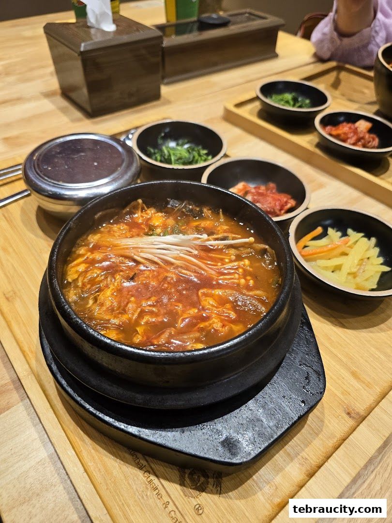A steaming bowl of Korean stew with side dishes on a wooden table.