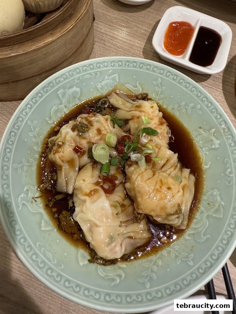 A close-up of wontons in a savory sauce, garnished with scallions and chili.