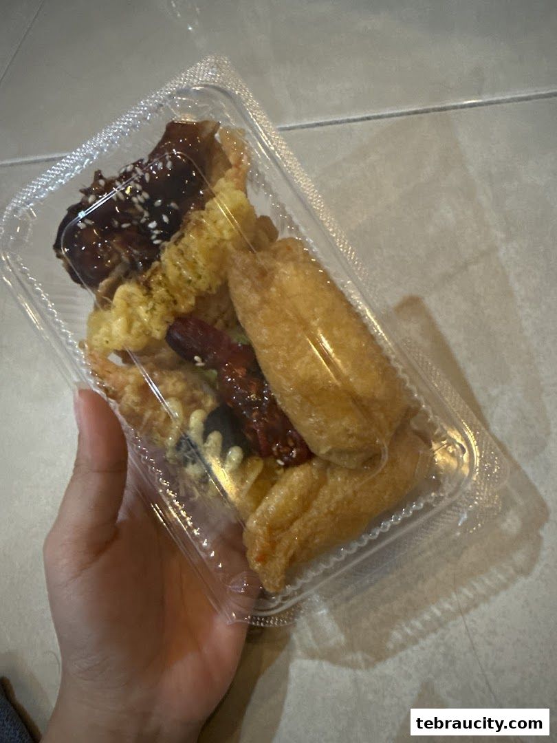 A hand holds a clear plastic container filled with assorted Japanese fried snacks.