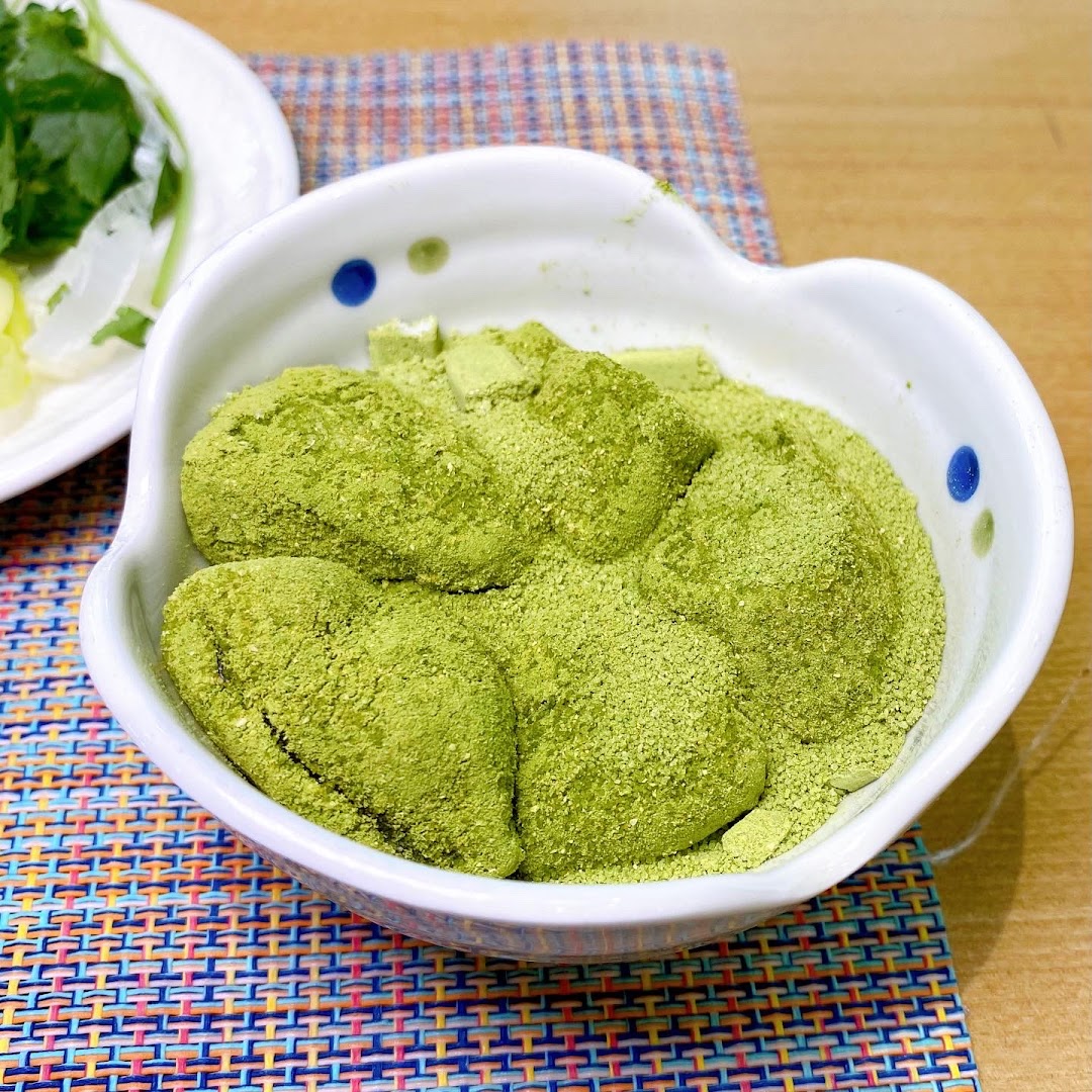 A bowl of green mochi-like desserts, generously coated with matcha powder, on a colorful placemat.