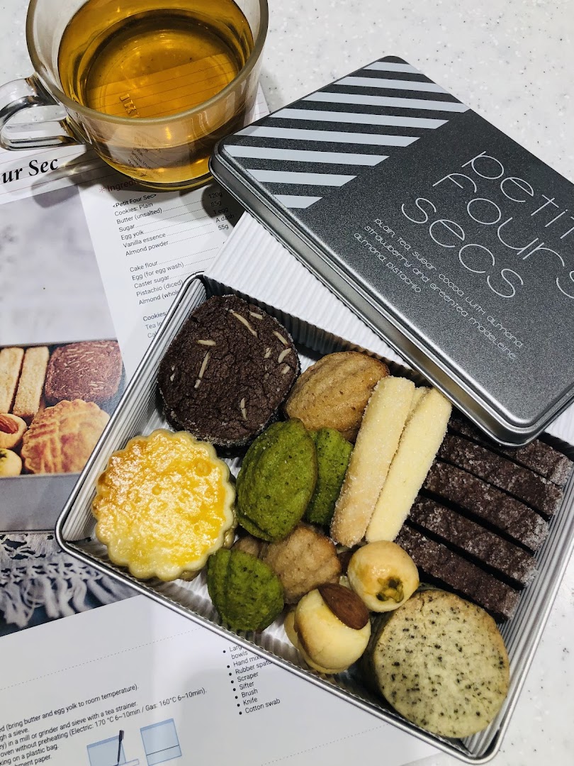 An open tin of assorted petit fours and cookies, with a cup of tea and a recipe sheet.