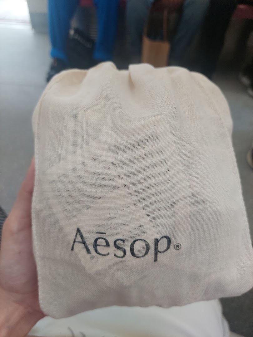 A hand holds a natural fabric drawstring bag featuring the Aesop logo, likely containing skincare products.