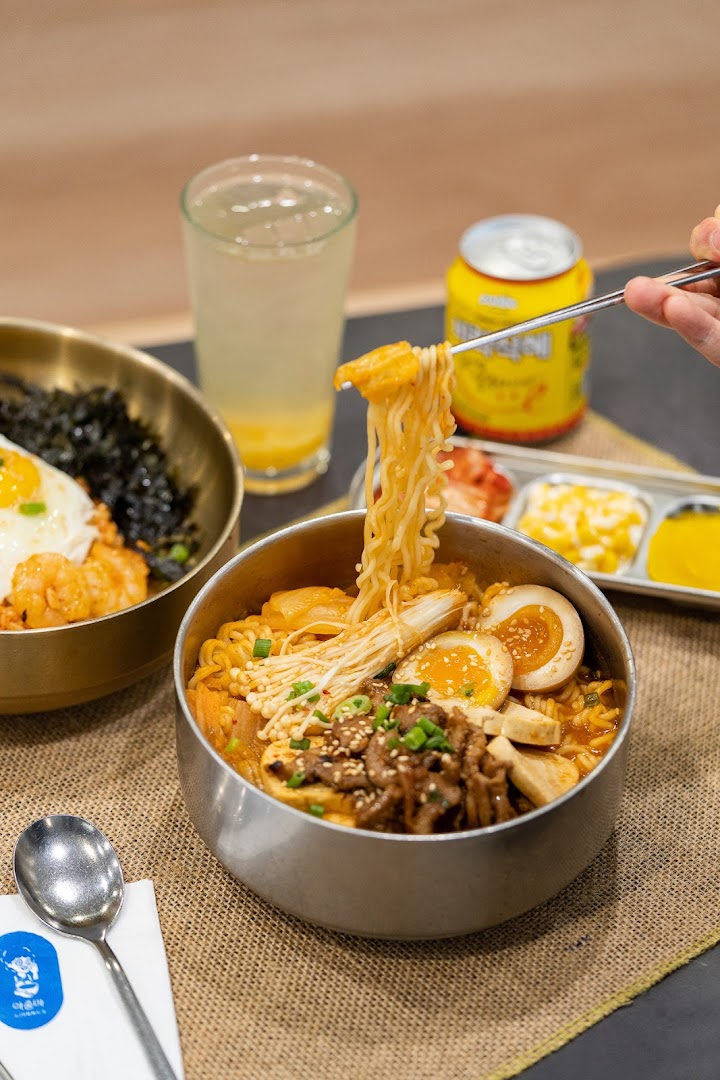 A close-up of delicious Korean ramen with various toppings, a rice dish, and drinks on a table.