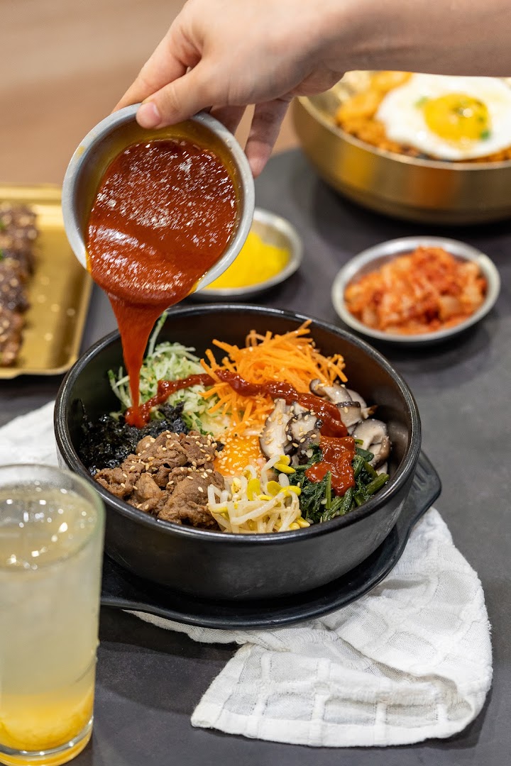 A hand pours spicy gochujang sauce over a vibrant bowl of bibimbap with various fresh ingredients.