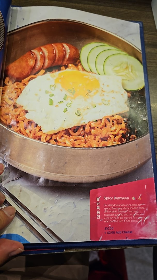 A menu page displaying a vibrant Spicy Ramyeon dish with sausage, egg, and cucumber.