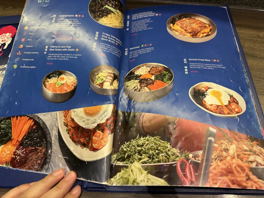 A vibrant menu displaying various Korean dishes like Jjajangmyeon, Kkal Guksu, Bibimbap, and Kimchi Fried Rice.