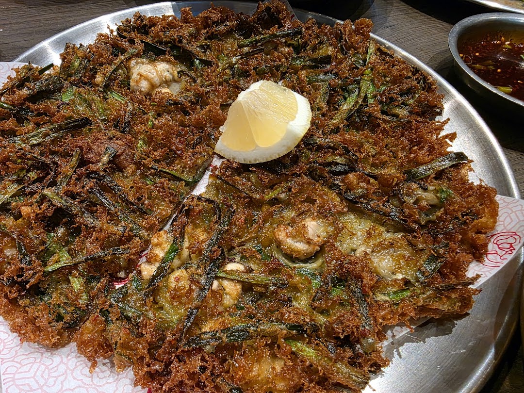 A close-up of a crispy Korean seafood and green onion pancake (Pajeon) with a lemon wedge on a metal plate.