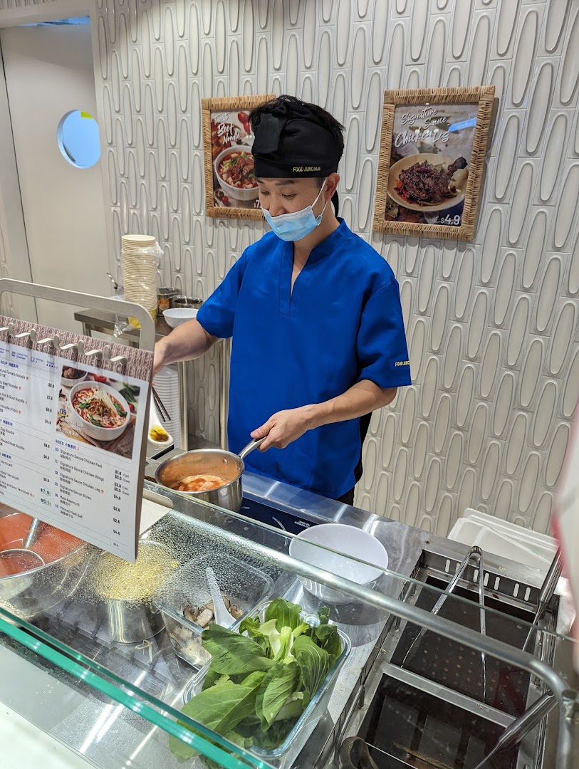 A chef in a blue uniform prepares a fresh noodle dish at the counter of Ali Tomato Soup Noodle.