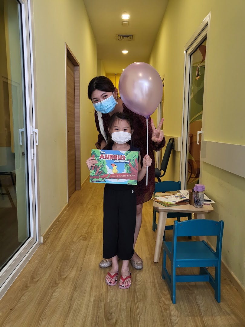 An adult and child, both masked, in a bright hallway, with the child holding an 'Aureus Adventure' book.