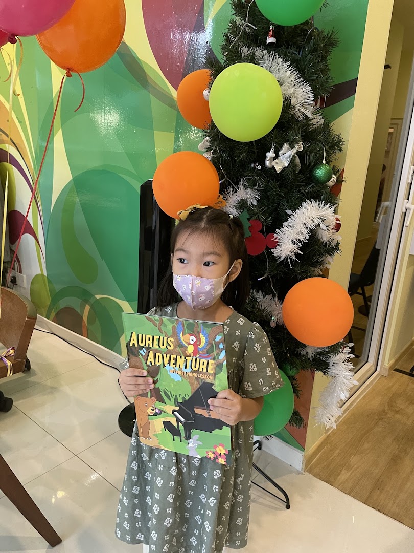 A young girl in a mask holds an 'Aureus Adventure' book next to a decorated Christmas tree and balloons.